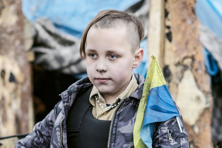 Ein Kind mit weißem Rosenkranz, ukrainischen Farben und Kampfweste. Zu jung um zu kämpfen, zu alt um nicht Teil eines historischen Ereignisses seines Landes zu werden, trägt es die traditionelle Haartracht der ukrainischen Kosaken. (Foto: Thomas Schell)